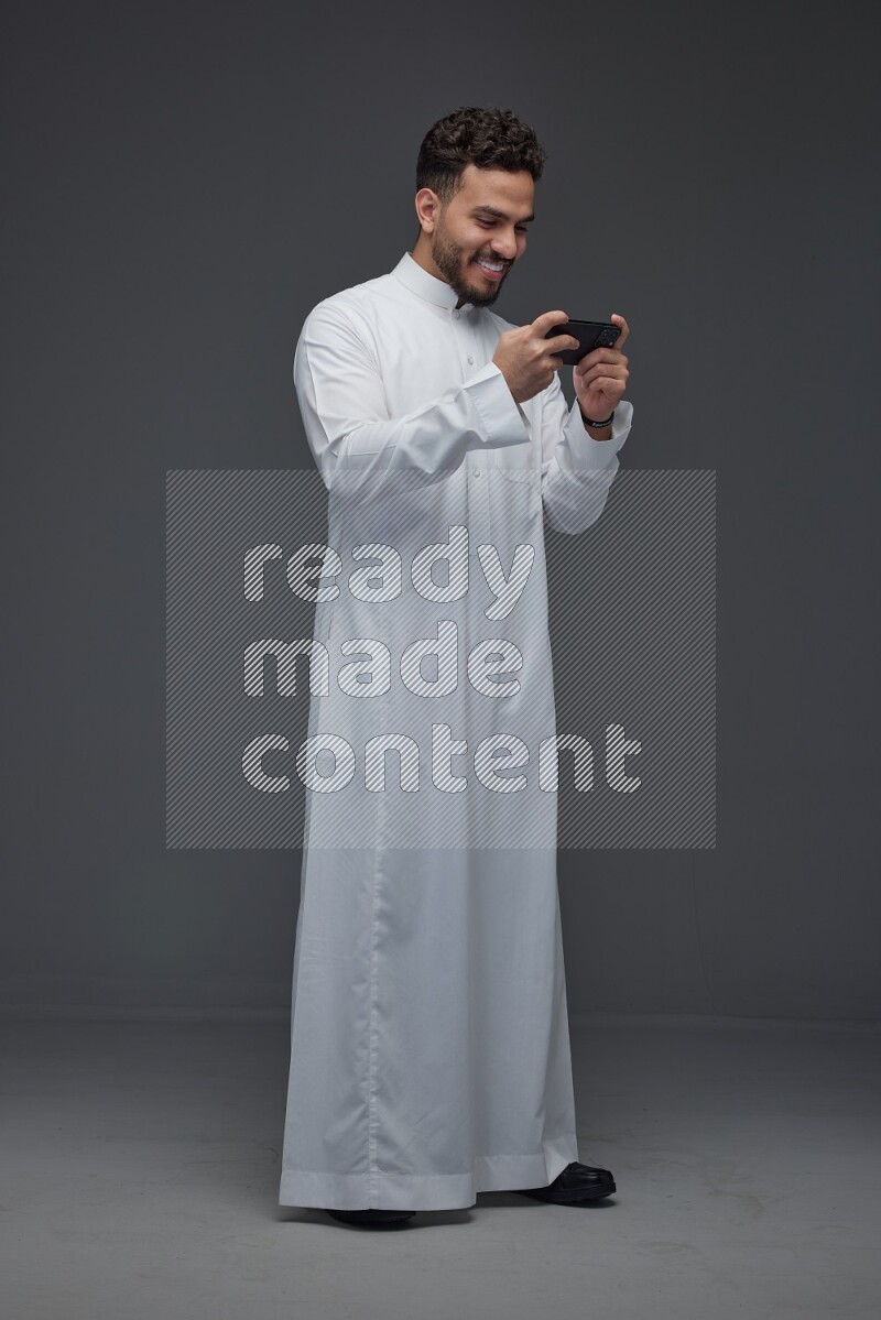 A Saudi man wearing Thobe and playing using his phone horizontally playing video games while standing and making different poses eye level on a gray background