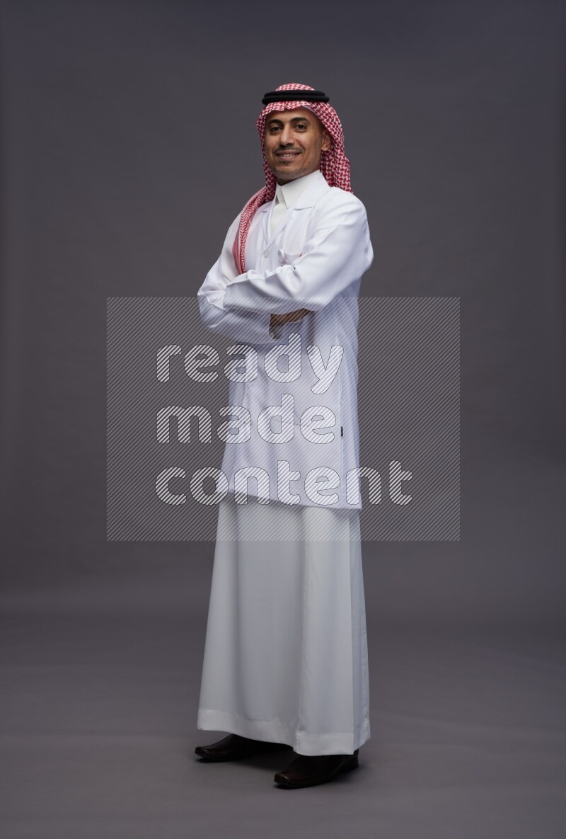 Saudi man wearing thob with lab coat and shomag with pocket employee badge standing with crossed arms on gray background