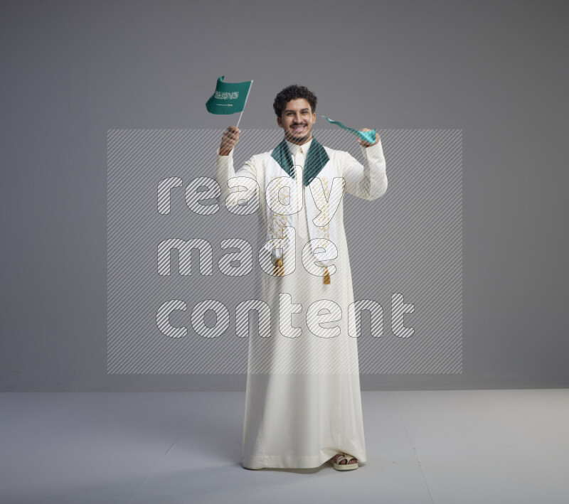 A Saudi man standing wearing thob and saudi flag scarf and holding small saudi flag on gray background