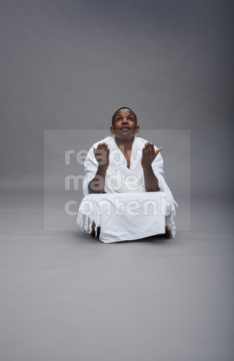 A man wearing Ehram sitting on floor dua'a on gray background