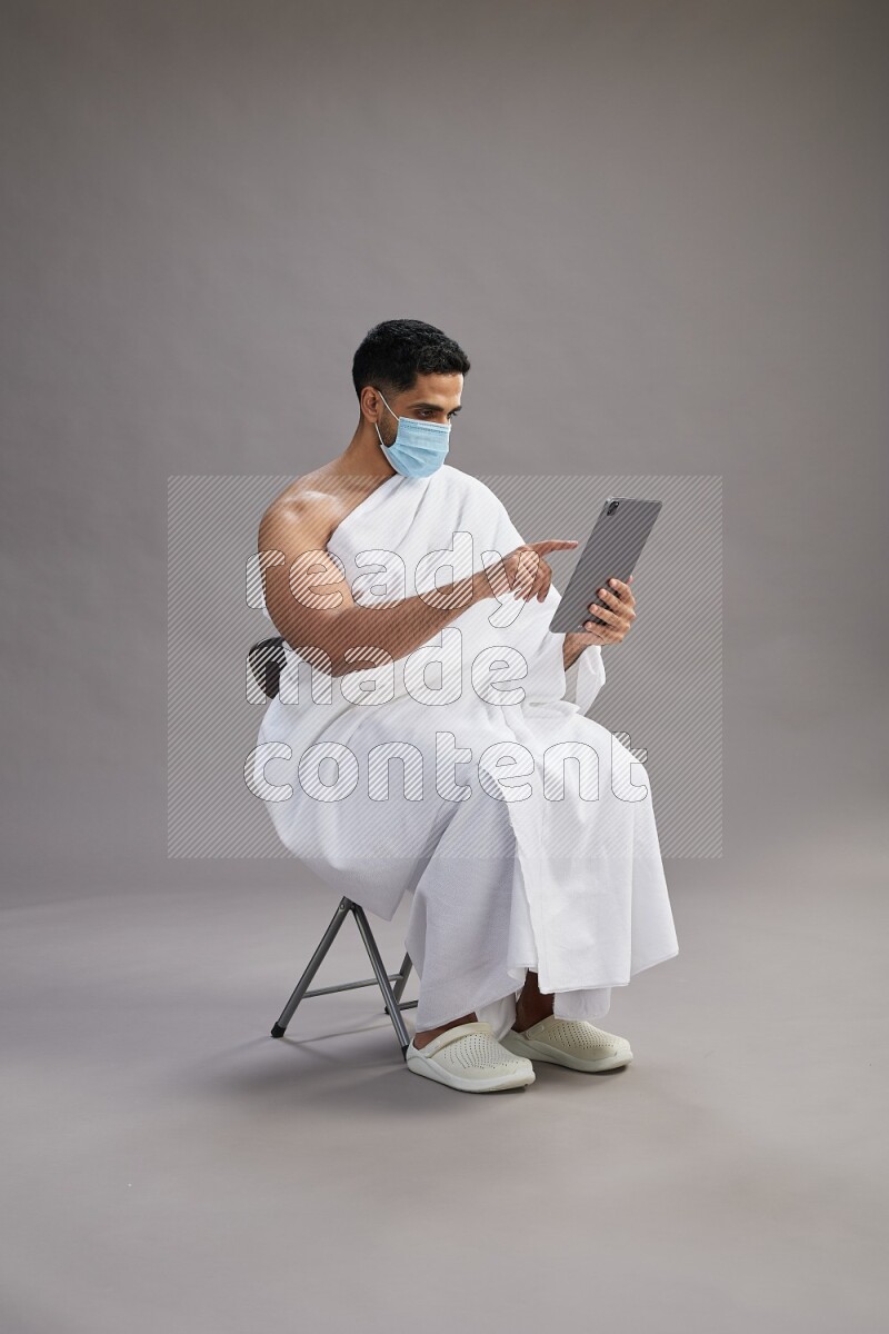 A man wearing Ehram with face mask sitting on chair holding a tablet on gray background