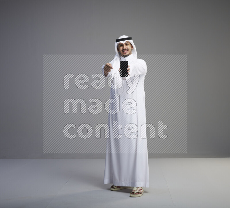 A Saudi man standing wearing thob and white shomag showing phone to camera on gray background