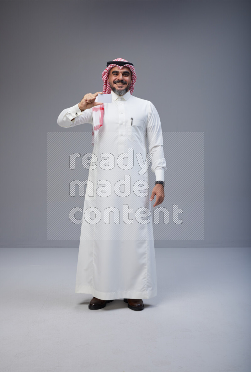 Saudi man Wearing Thob and red Shomag standing holding ATM card on Gray background