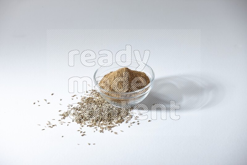 A glass bowl full of cumin powder with cumin seeds beneath it on white flooring