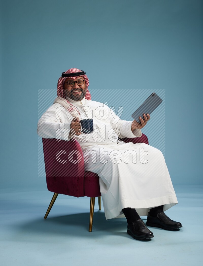 Saudi Man with shimag sitting on chair working on tablet on blue background