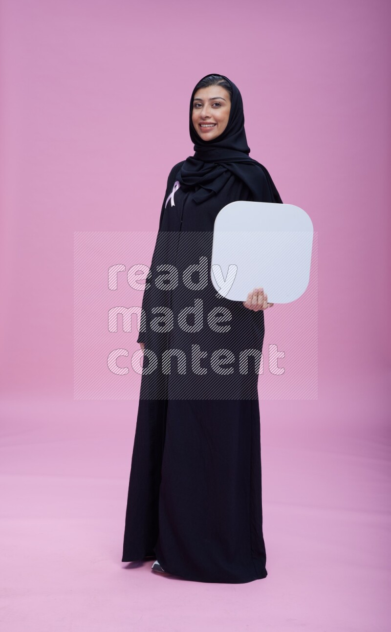 Saudi woman wearing pink ribbon on Abaya standing holding social media signs on pink background