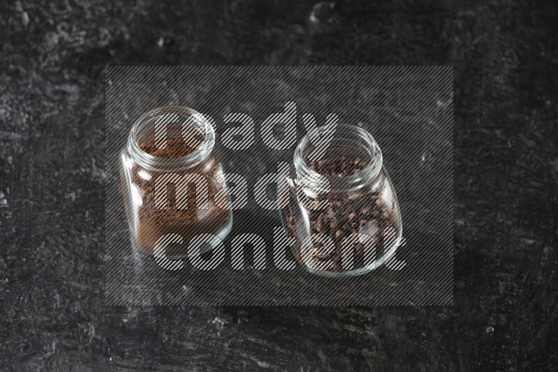 Glass spice jars full of cloves and cloves powder on textured black flooring