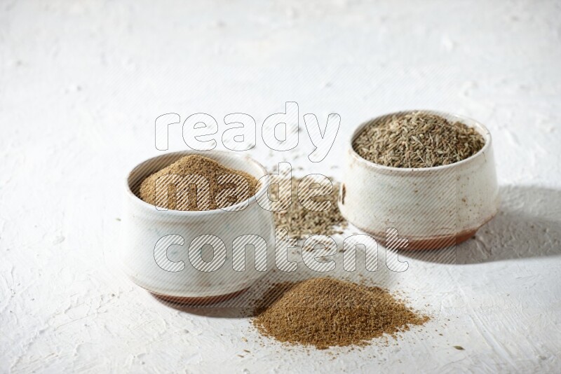2 beige bowls full of cumin seeds and powder with spilled powder and seeds on textured white flooring