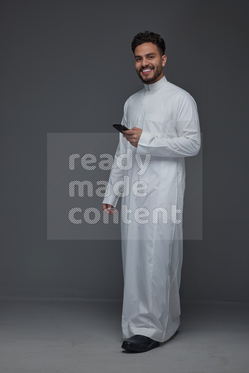 A Saudi man wearing Thobe standing and using his phone eye level on a gray background