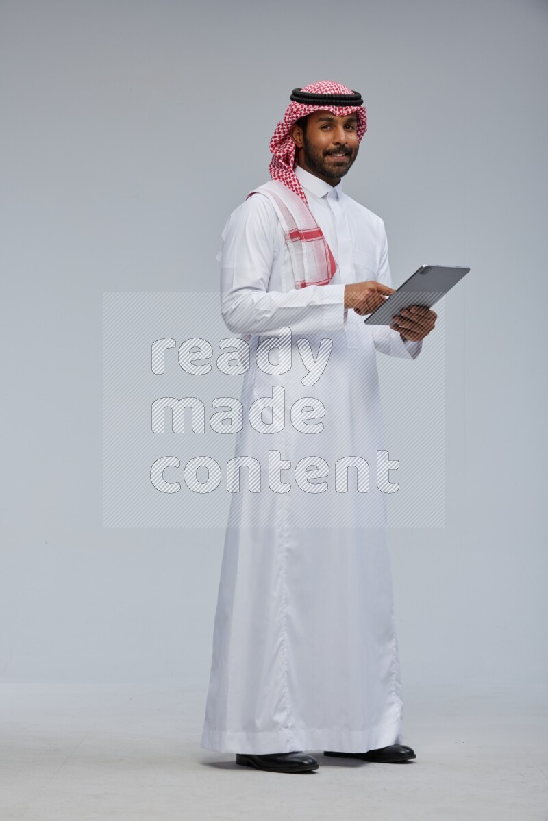 Saudi man Wearing Thob and shomag standing working on tablet on Gray background