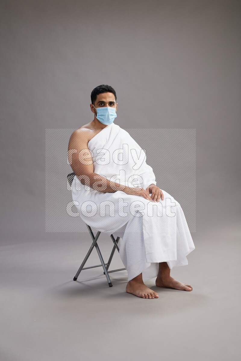 A man wearing Ehram with face mask sitting on chair Interacting with the camera on gray background