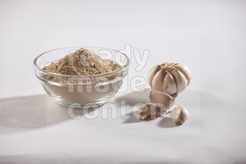 A glass bowl full of garlic powder with garlic bulb and some cloves beside it on a white flooring