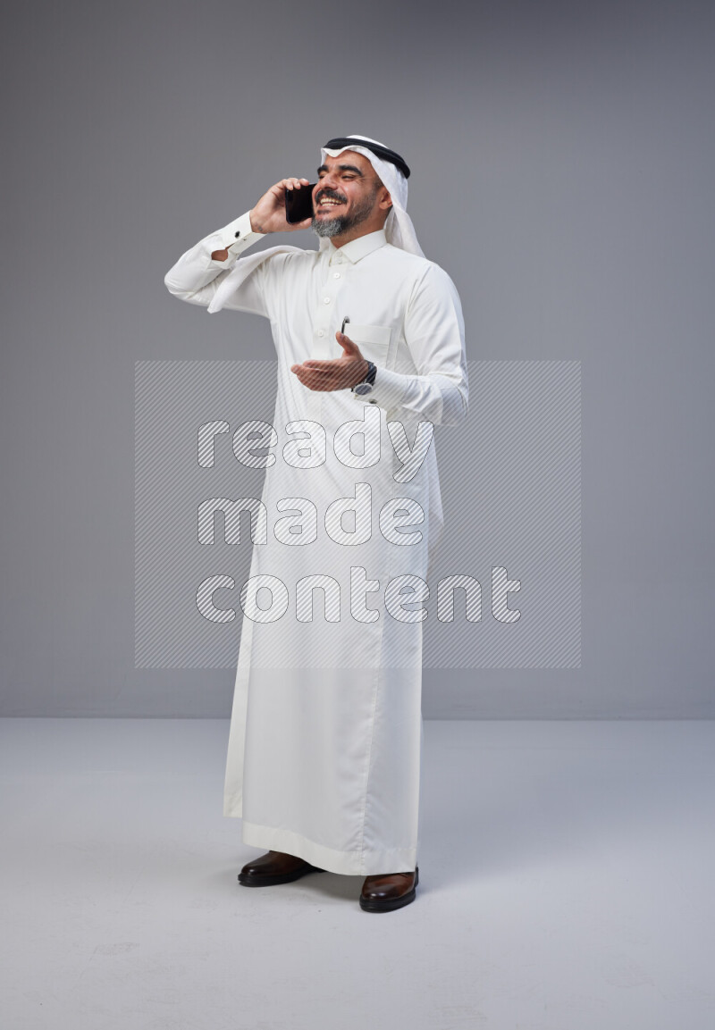 Saudi man Wearing Thob and white Shomag standing talking on phone on Gray background