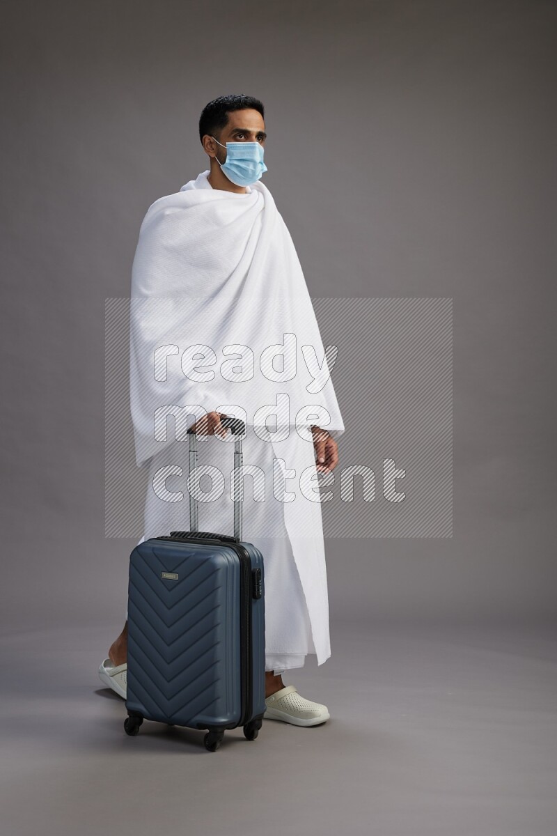A man wearing Ehram with face mask Standing holding traveling bag on gray background