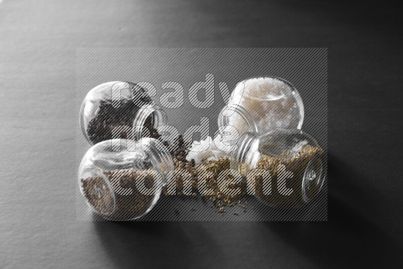 4 glass spice jars full of salt, black peppers, white peppers and cumin on black flooring