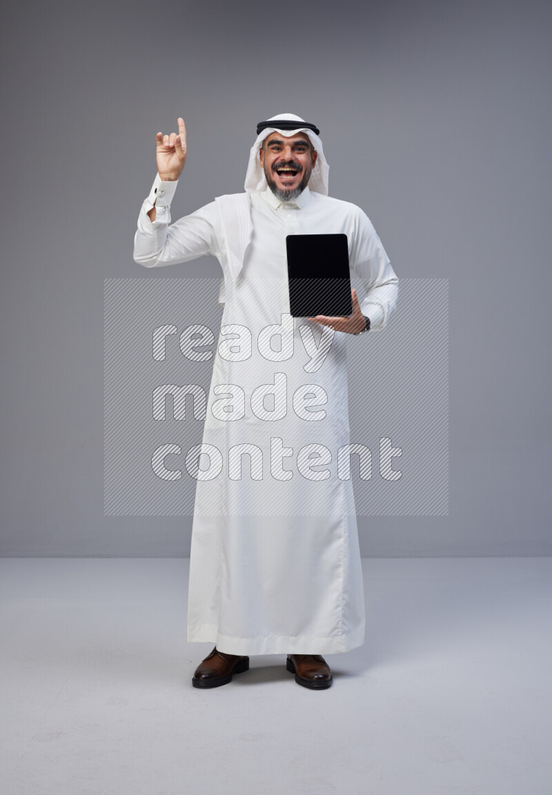 Saudi man Wearing Thob and white Shomag standing showing tablet to camera on Gray background