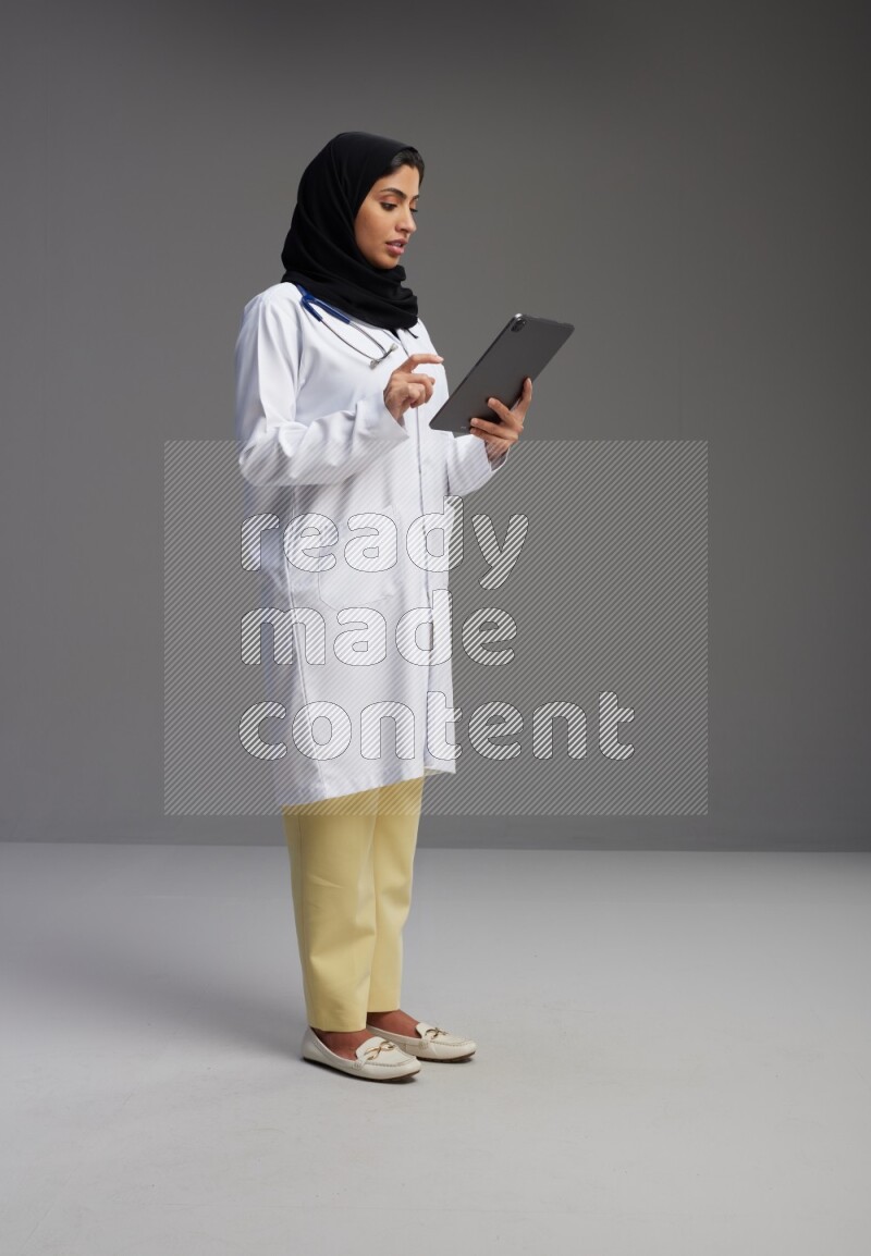 Saudi woman wearing lab coat with stethoscope standing working on tablet on Gray background