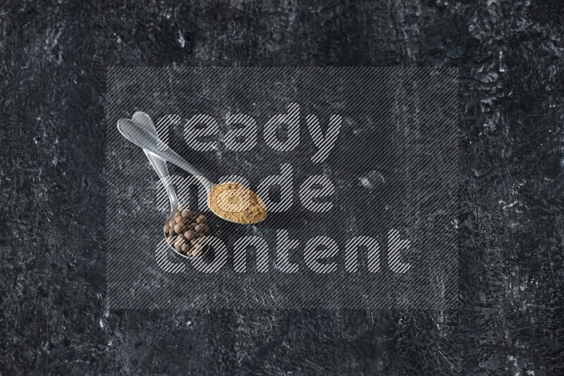 2 metal spoons full of allspice powder and whole balls on a textured black flooring