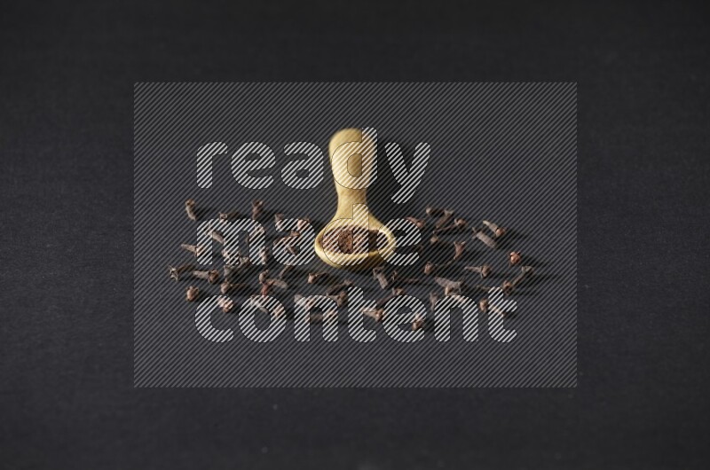 A wooden spoon full of cloves powder with spreaded whole cloves on a black flooring