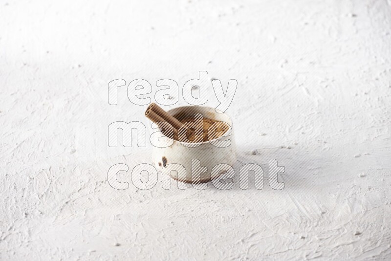 Ceramic beige bowl full of cinnamon powder with a cinnamon stick on a textured white background