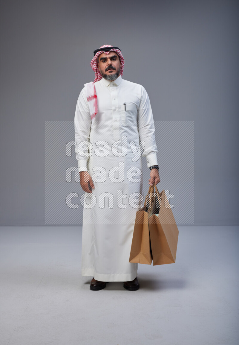 Saudi man Wearing Thob and red Shomag standing holding shopping bag on Gray background