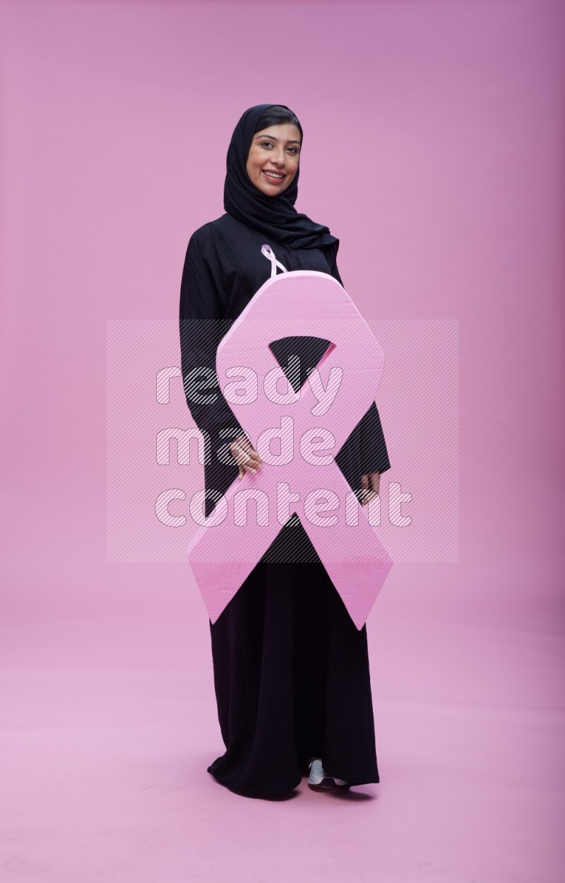 Saudi woman wearing pink ribbon on abaya standing holding awareness ribbon on plain pink background