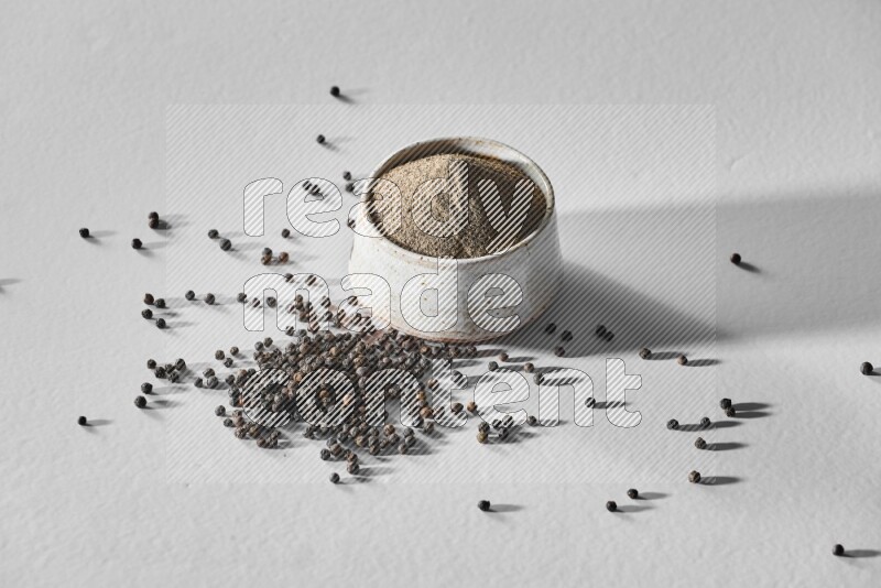 فلفل أسود مطحون في وعاء أبيض من السيراميك مع حبوب فلفل أسود علي خلفية بيضاء