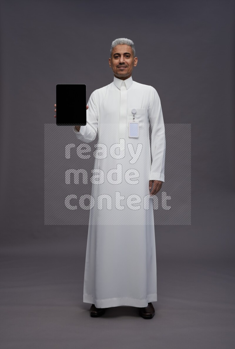 Saudi man wearing thob with pocket employee badge standing showing tablet to camera on gray background