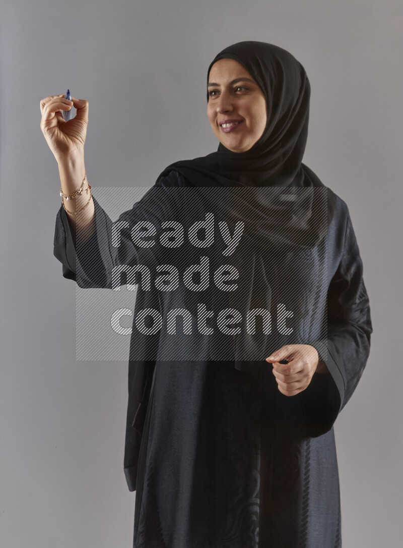 Woman in black abaya and hijab holding a marker pen to write on a transparent board with different reactions and expressions on grey background