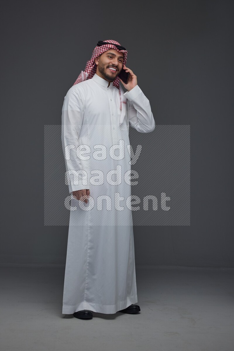 A Saudi man wearing Thobe and Shmagh talking in the phone eye level on a gray background