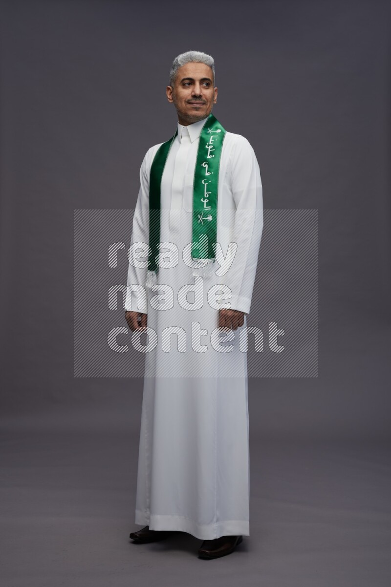 Saudi man wearing thob standing holding Saudi flag on gray background