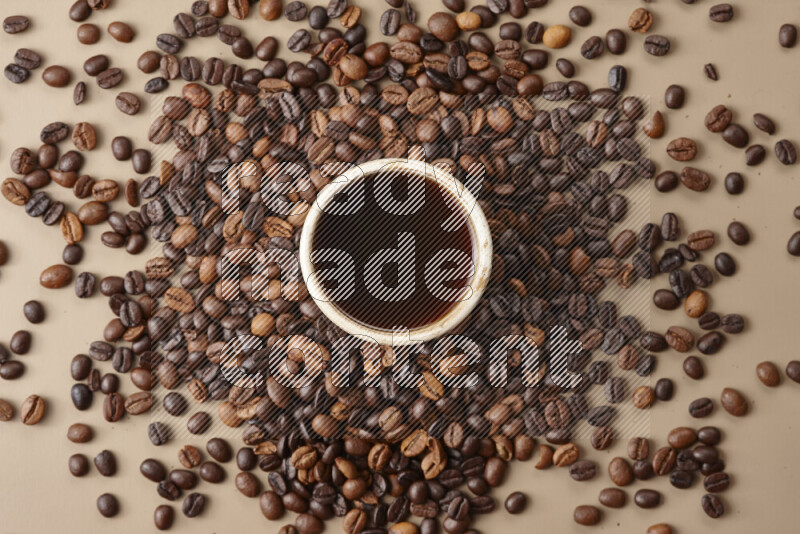 A beige pottery cup of coffee surrounded by roasted coffee beans on beige background