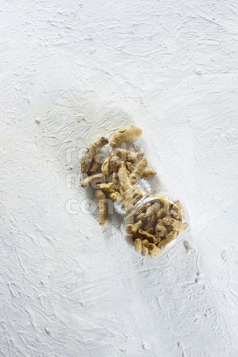 A flipped glass spice jar full of dried turmeric fingers and fell out it on textured white flooring