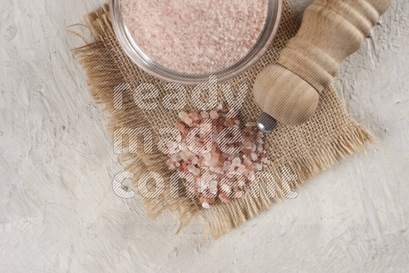 A glass bowl full of pink himalayan salt with a wooden grinder on a burlap fabric all on white background