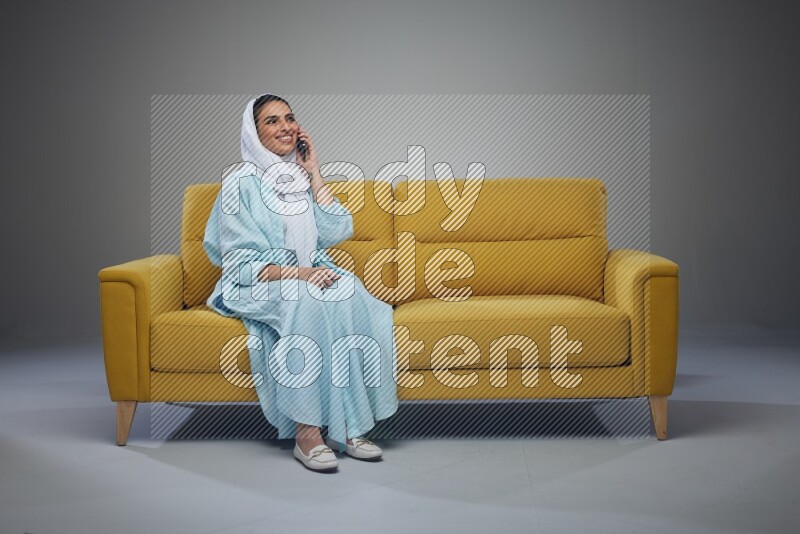 A Saudi woman wearing a light blue Abaya and a white head scarf sitting on a yellow sofa and talking in the phone while making different poses eye level on a grey background