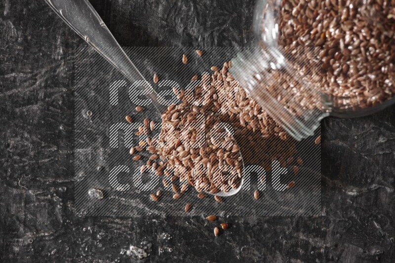 A glass spice jar full of flaxseeds flipped and seeds spread out with a metal spoon full of the seeds on a textured black flooring