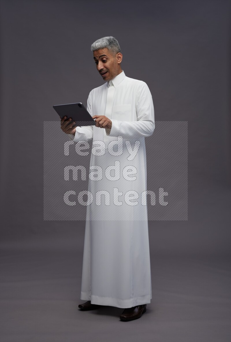 Saudi man wearing thob standing working on tablet on gray background
