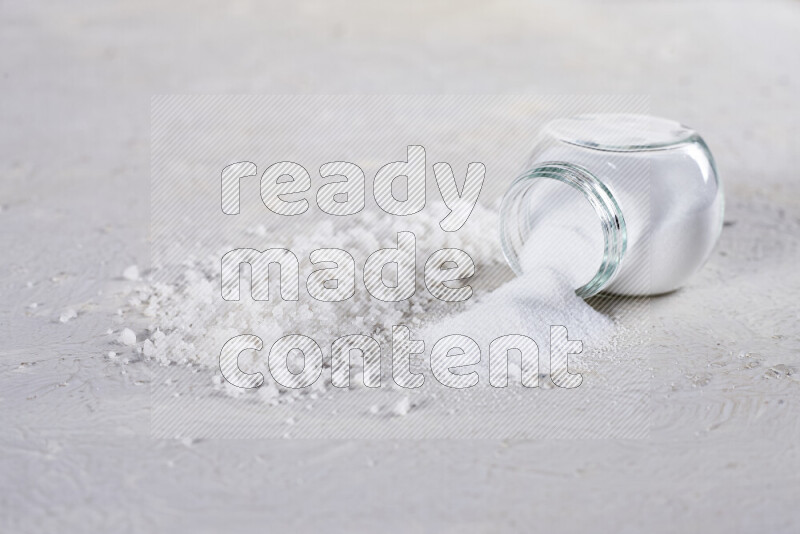 A glass jar full of table salt with some sea salt crystals beside it on a white background
