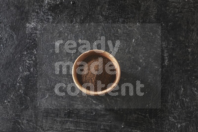 A wooden bowl full of cloves powder on a textured black flooring