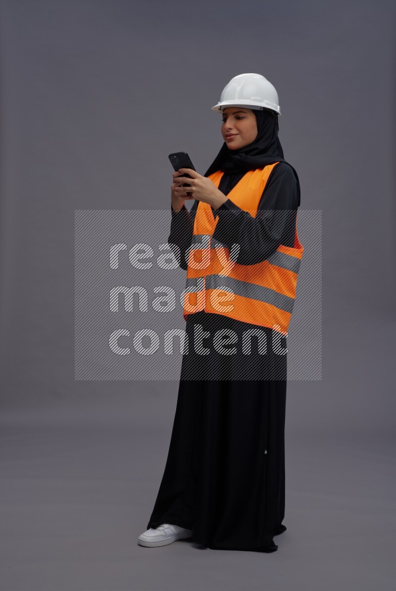 Saudi woman wearing Abaya with engineer vest standing texting on phone on gray background