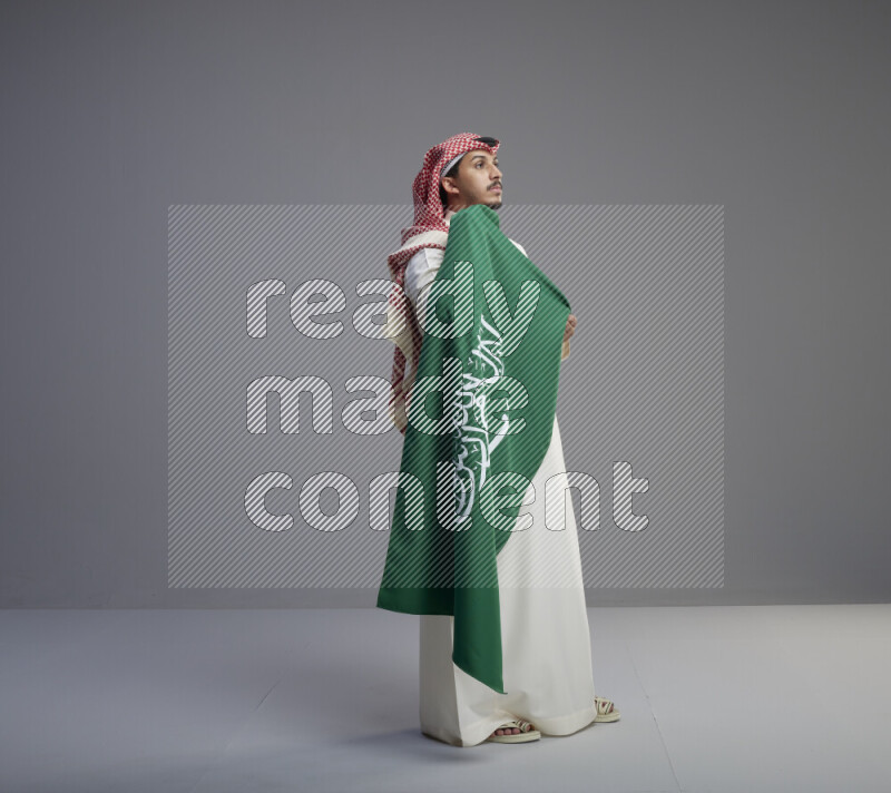 A saudi man standing wearing thob and red shomag holding big saudi flag on gray background