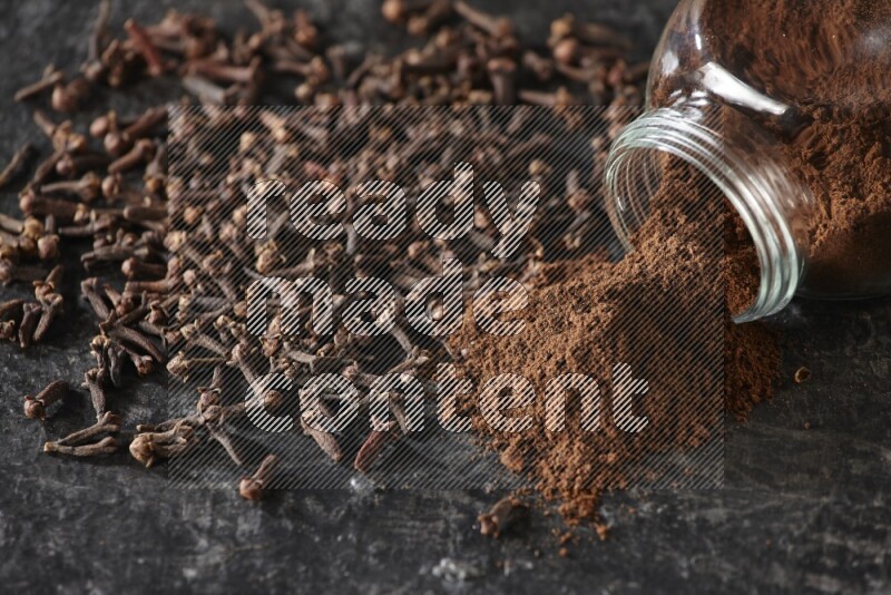 A flipped glass spice jar full of cloves powder and powder came out of it with cloves spread on textured black flooring