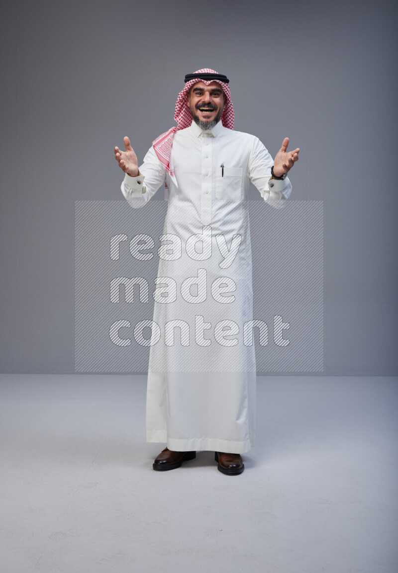 Saudi man Wearing Thob and red white Shomag standing interacting with the camera on Gray background