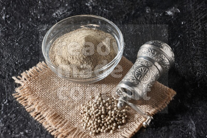 A glass bowl full of white pepper powder with white pepper beads on a burlap piece of fabric and a metal grinder on textured black flooring