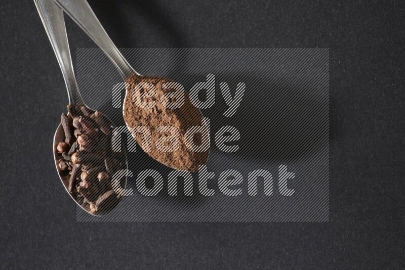 2 metal spoons, one full of cloves grains and the other full of cloves powder on a black flooring