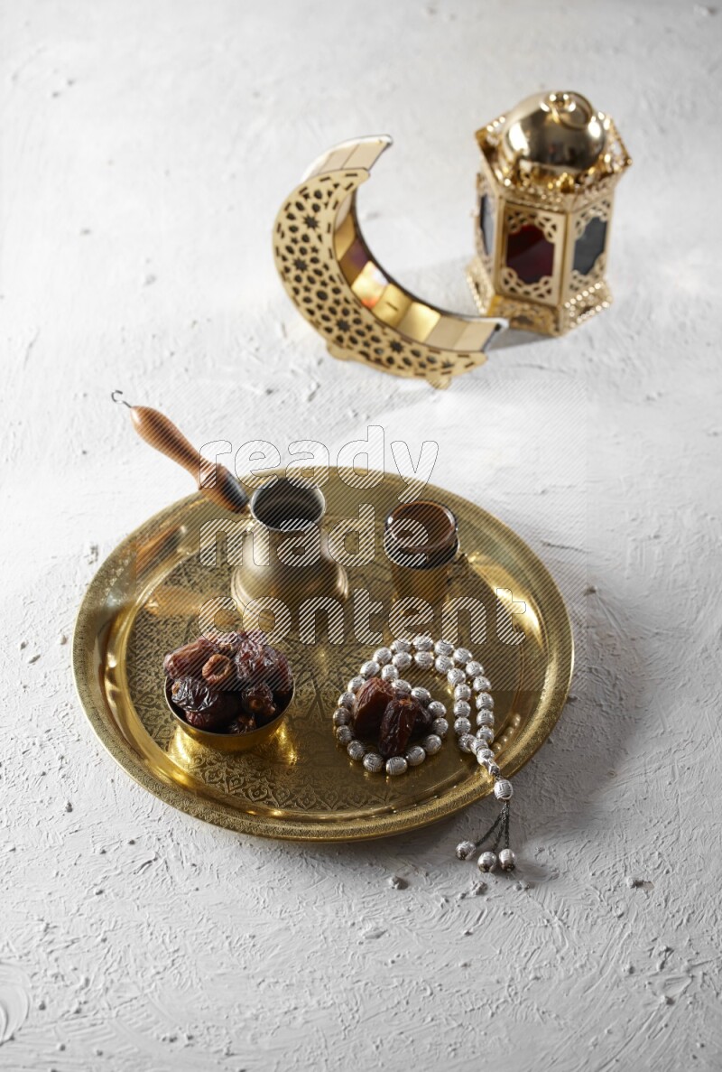 Dates in a metal bowl with coffee and prayer beads on a tray beside lanterns in a light setup
