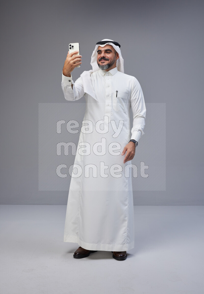 Saudi man Wearing Thob and white Shomag standing taking selfie on Gray background