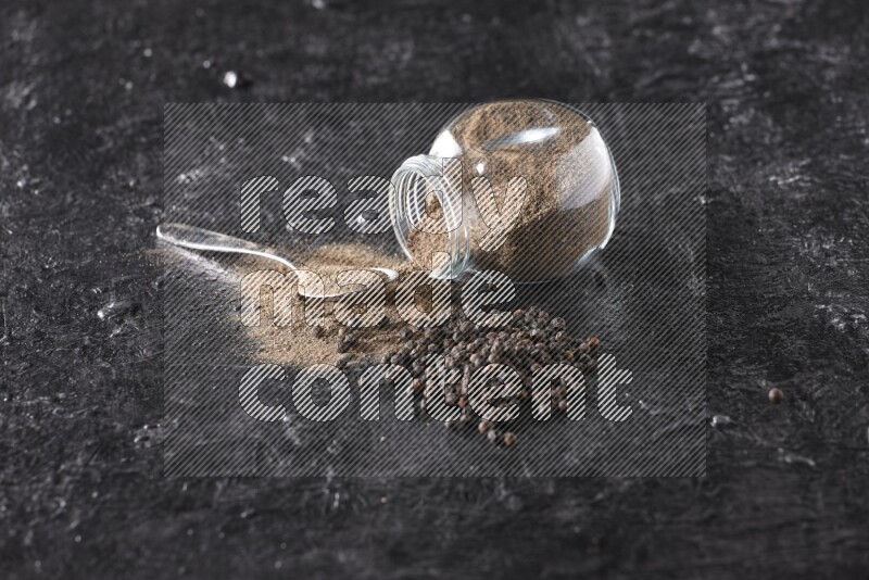 A flipped glass spice jar full of black pepper powder with a metal spoon full of powder and pepper beads on textured black flooring