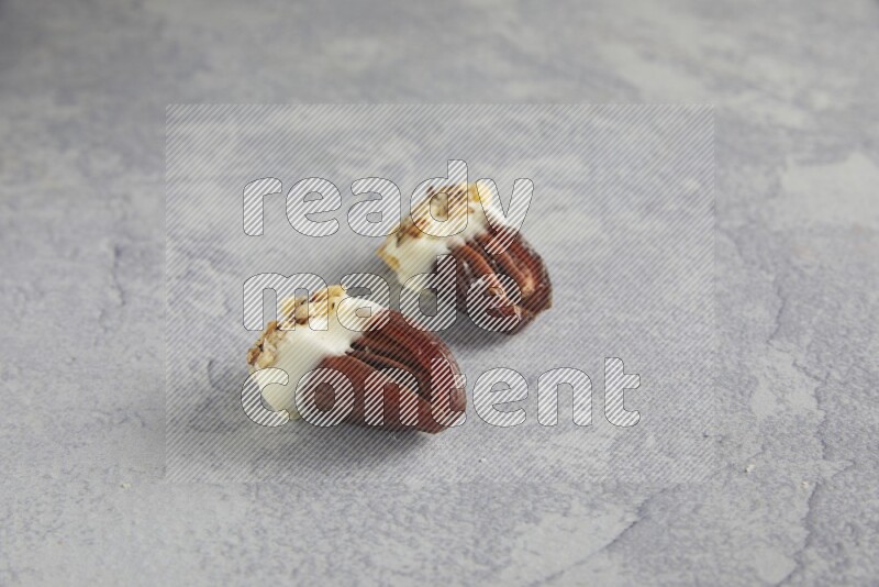 Two pecan stuffed date covered with white chocolate and chopped walnuts on alight grey background