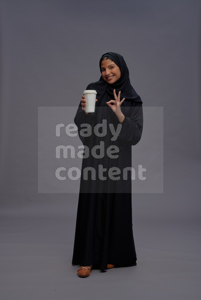 Saudi woman wearing Abaya standing holding paper cup on gray background